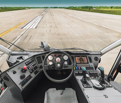 The inside of an airport fire truck cab shows the centered seat, wheel, controls and the large window with a runway visible into the distance. 