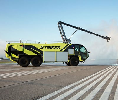 A yellow and black Striker ARFF vehicle is on a runway with white striping spraying water into the distance.  