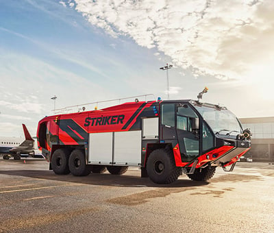 A red and white airport fire truck with ‘Striker’ in black letters on the side is parked at an airport with a terminal building and aircraft visible in the background. 