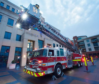 A red and white fire truck is positioned in front of a 4-story building with the aerial platform raised and scene lights turned on. 