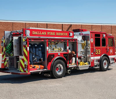 A red municipal fire truck is parked in front of a red brick building with all compartments open to show the compartment space.  