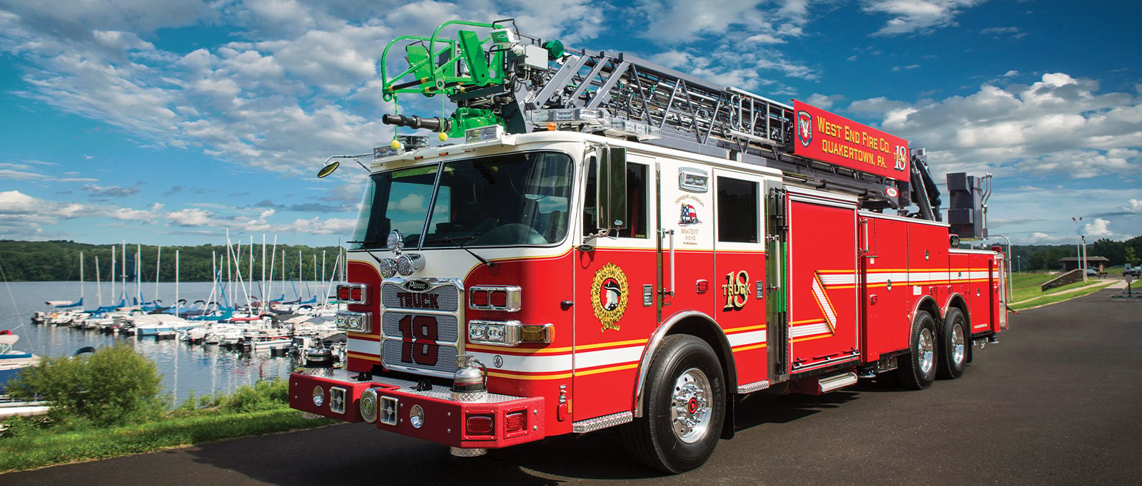 A red aerial ladder fire truck is parked on an asphalt road in front of a marina with a blue cloud-filled sky.