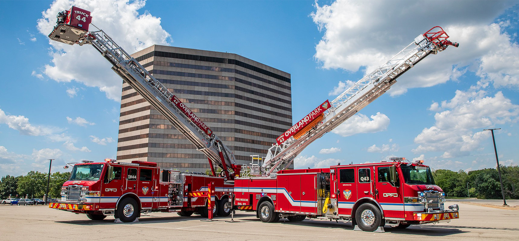 Two types of aerial fire trucks are pictured with raised ladders in front of a large office complex. 