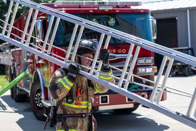 A firefighter carries an aluminum ground ladder with a red fire truck in the background. 