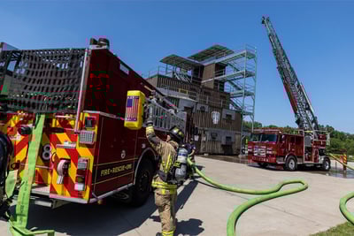A fire scene shows a firefighter pulling a ground ladder off the side of a red apparatus with a high-rise building and deployed aerial apparatus on a fire truck in the background. 