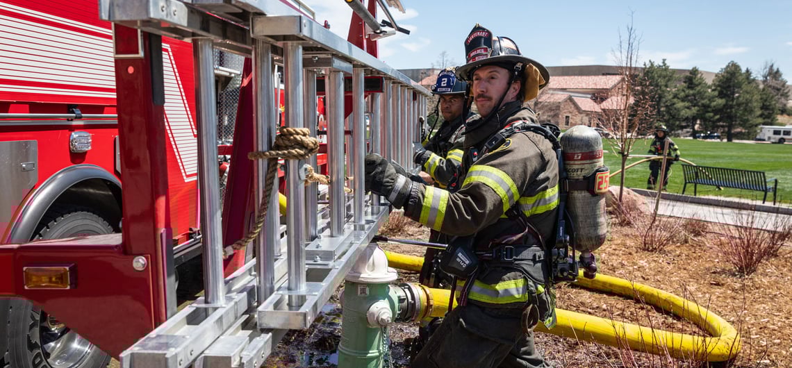 Two firefighters work together to remove an aluminum ground ladder from an external fire truck ladder rack outside in a city park area. 