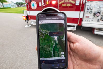 A hand is holding a cell phone that shows fire truck collision avoidance technology on the screen with a fire engine in the background.  