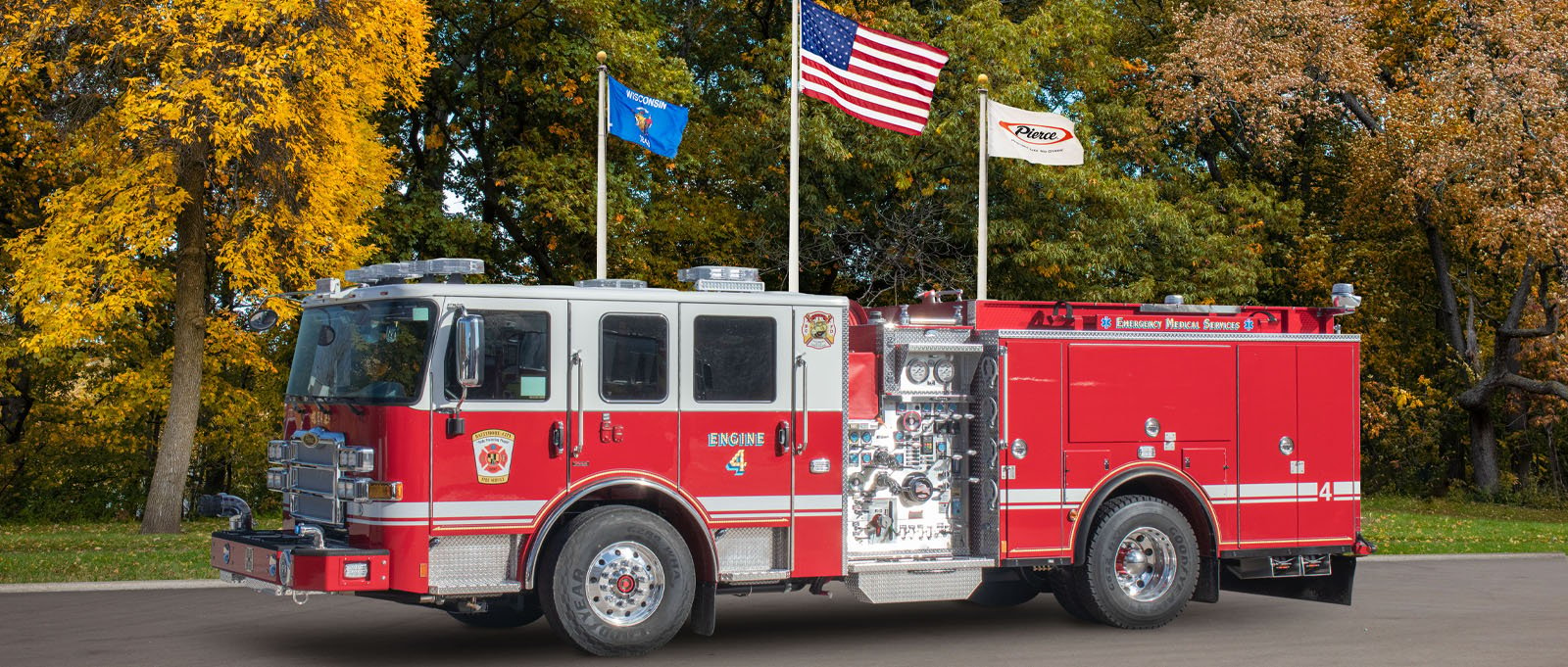 A red and white Baltimore City Fire Department engine is parked with trees and flags in the background. 