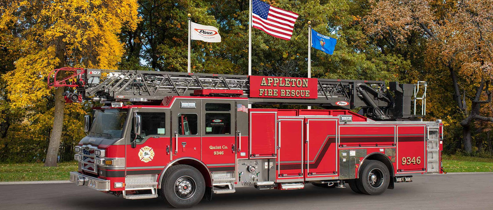 A red Appleton Fire Department aerial fire truck is parked with flags and a fall-colored tree line in the background. 