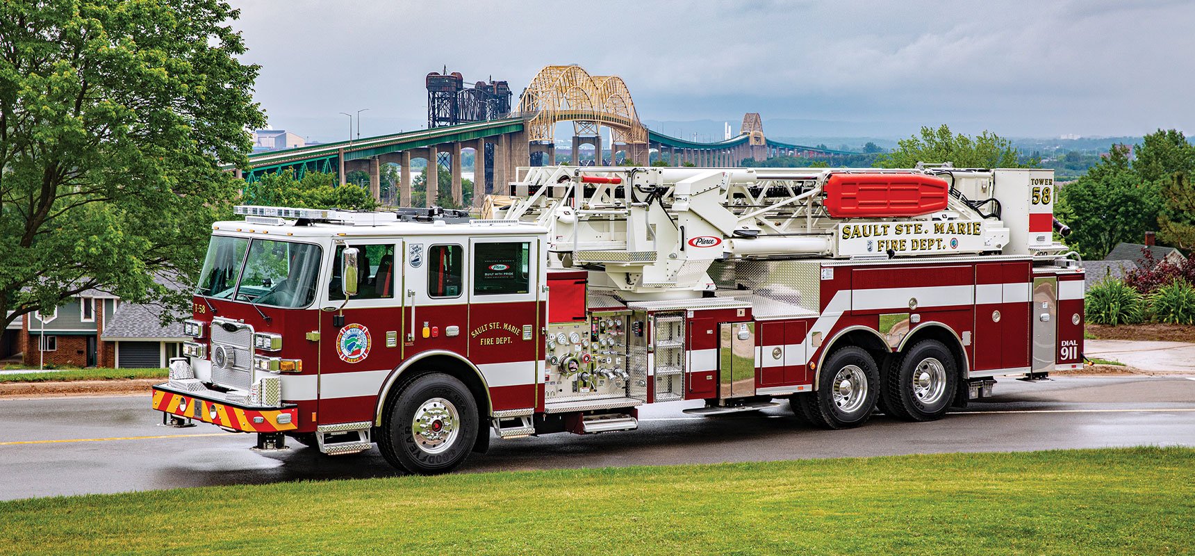 A red and white urban aerial fire truck is parked in front of a large bridge and green landscape.