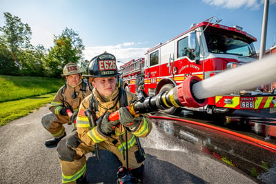 Two urban firefighters are kneeling on pavement holding a fire nozzle as it sprays water in front of a parked urban fire truck.