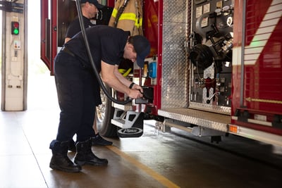 A red electric fire truck is parked in a fire station bay and is being connected to the charging station by a firefighter.  