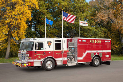 A red pumper fire apparatus on street with three flags and green and yellow colored trees in the background. 