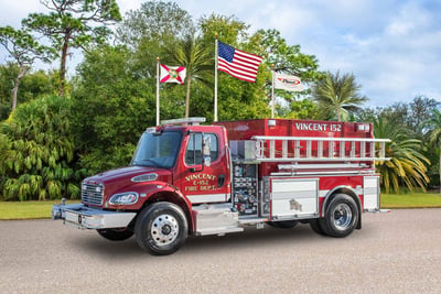 A red tanker fire apparatus on street with three flags green trees in the background 