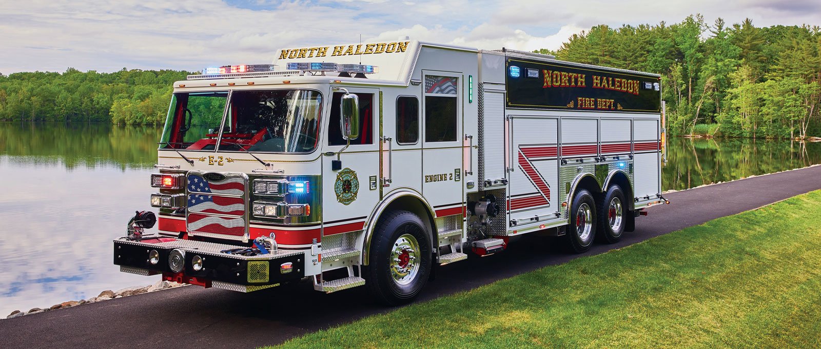 A white tanker fire apparatus sitting on a road alongside a lake with green grass and trees in the background. 