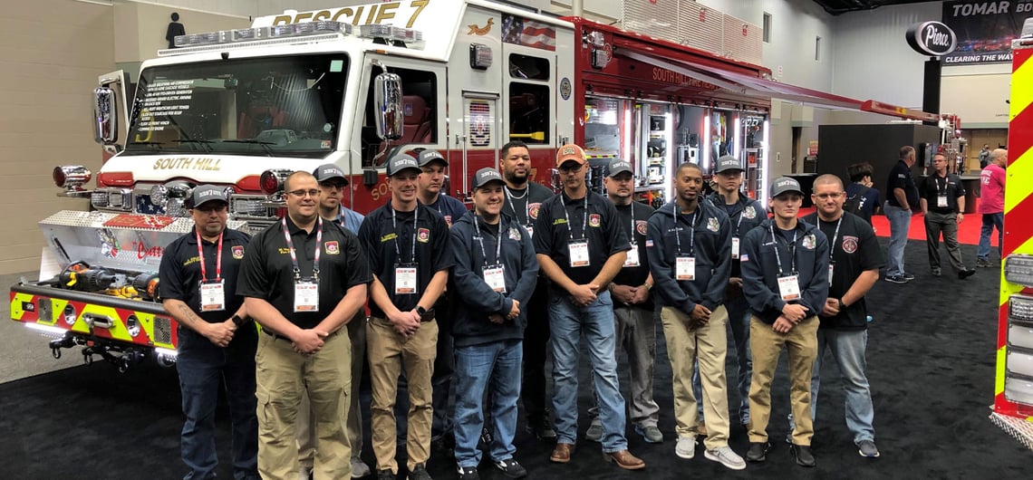 A group of firefighters in front of a red fire apparatus on a black carpet in a convention center hall.