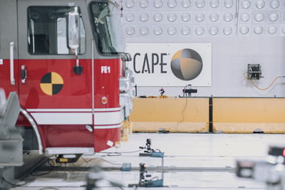 A close-up image of a red fire truck cab with an orange and black circle on the passenger door, which indicates the cab is being used for crash testing. A sign with the location of the testing, CAPE, is visible in the background.