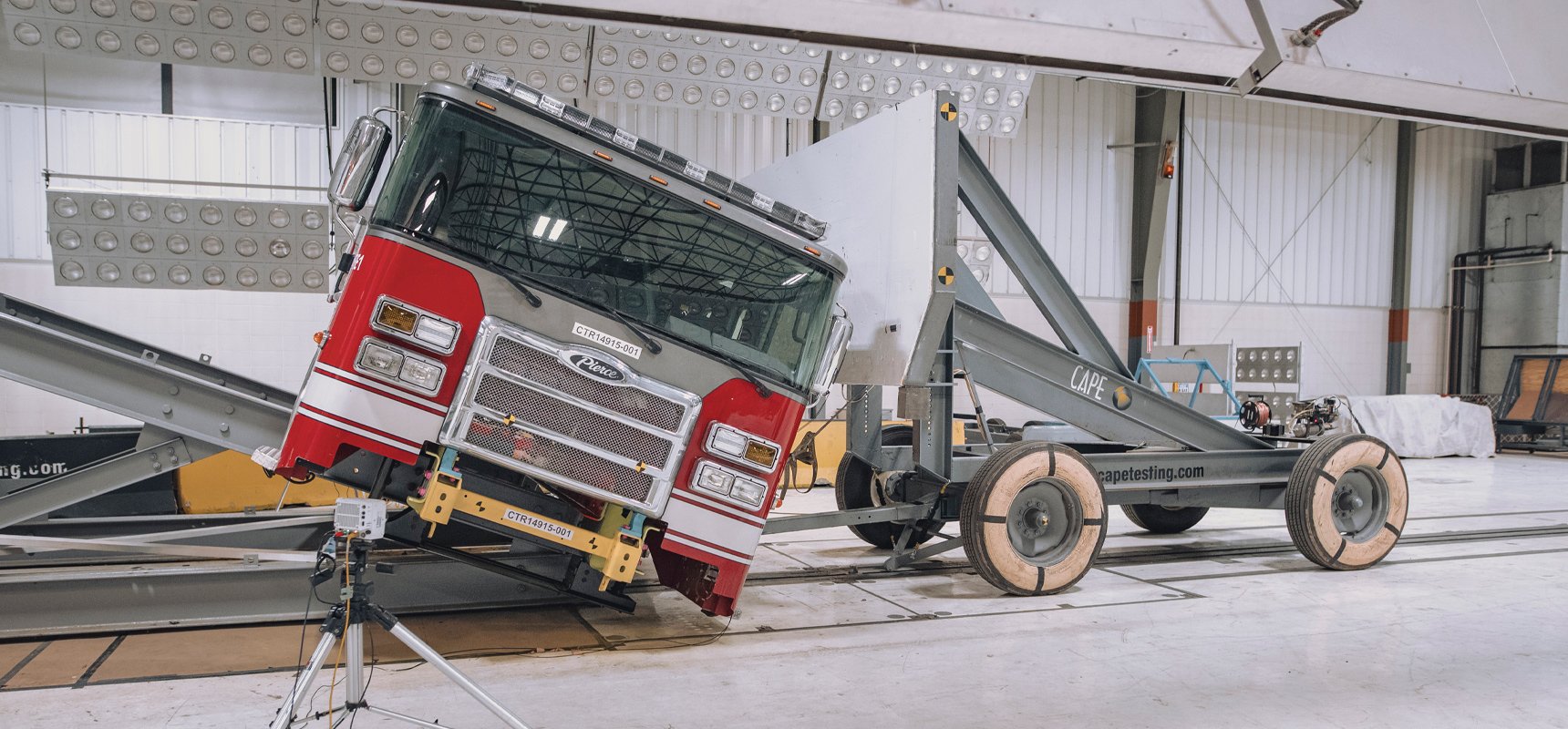 A tilted red Pierce fire truck cab is being hit with a large barrier to simulate a fire truck rollover crash scenario in a fire truck cab crash testing facility.