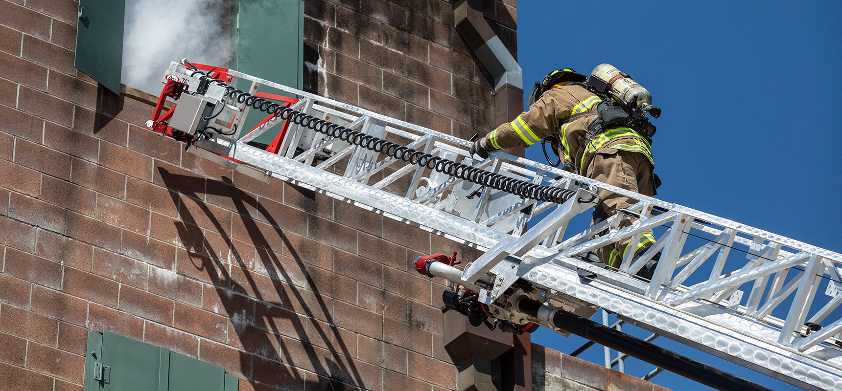 A firefighter in full turnout gear ascents an aerial ladder device toward an open window as smoke billows out. 