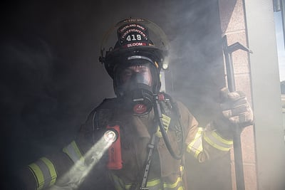 A firefighter holds a pick ax and dons full protective gear and SBCA mask in a smoke-filled structure.