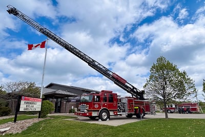 A red Pierce rear-mount aerial apparatus is positioned outside the Guelph Fire Department in Ontario, Canada, with the ladder extended overhead and the department in the background. 