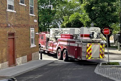 A red Pierce mid-mount aerial fire truck is pictured driving down a very narrow street and turning left at a red ‘Arret’ sign in Quebec, Canada. 