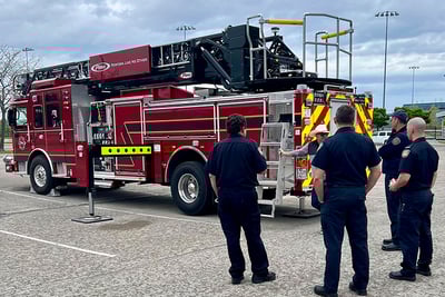 Five firefighters stand near the rear end of a red Pierce rear-mount aerial apparatus talking as the truck sits parked in a large lot outdoors. 
