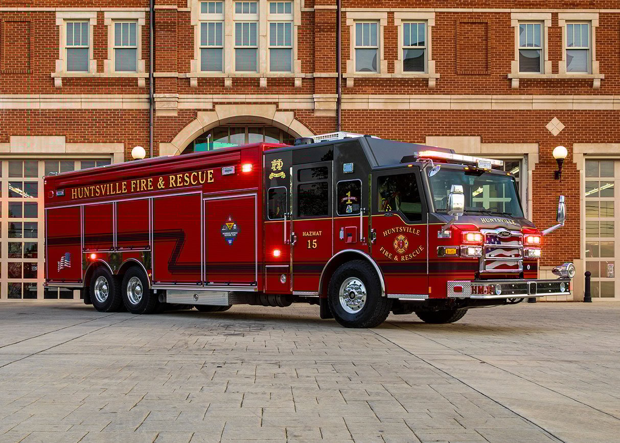 The front officer's side of a combination heavy-duty rescue fire truck in front of a brick fire station.