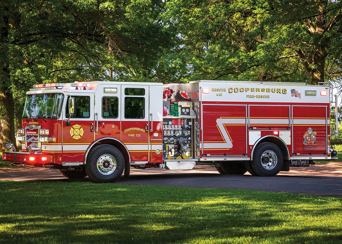 The officer's side of a Heavy-duty rescue pumper with the lights on parked in the shade under green trees.