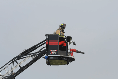 A volunteer fire fighter in full gear, stands in the bucket of a 100' ladder Pierce fire truck.