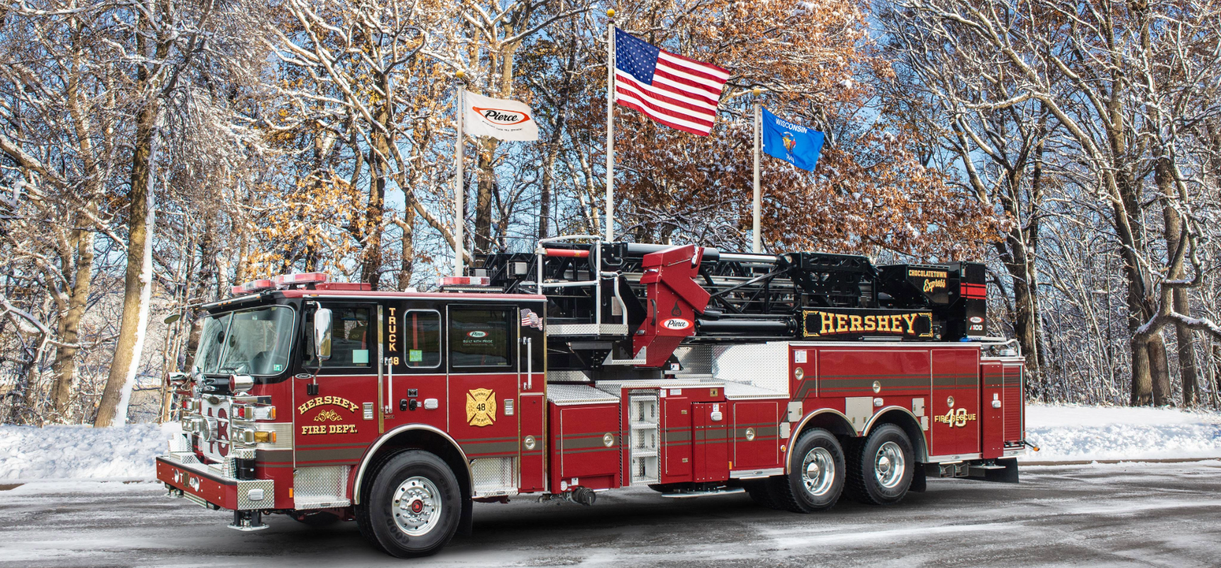 A red and black Pierce Enforcer pumper sits on a snowy road with three flagpoles and snow-covered trees in the background.