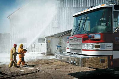 Firefighters holding hose while putting out a fire next to a Pierce Manufacturing fire truck that has corrosion and chemical resistance properties.