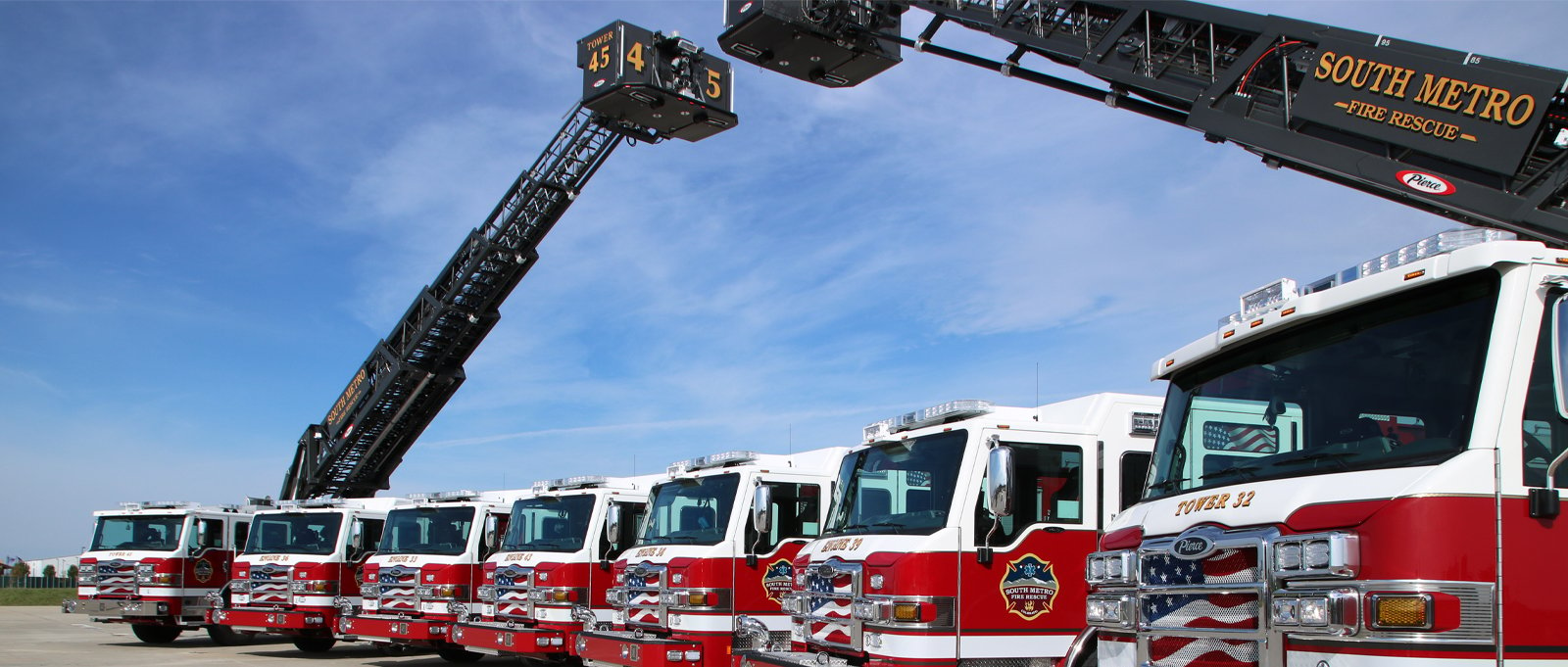 Seven Pierce fire apparatus lined up displaying their aerial ladders on the ends extended over the apparatus in the middle showing a full Pierce standardized fleet.