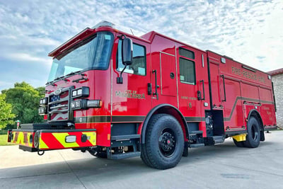 A red PUC fire truck is parked on an angle  outside with a green trees and a blue sky with clouds in the background.