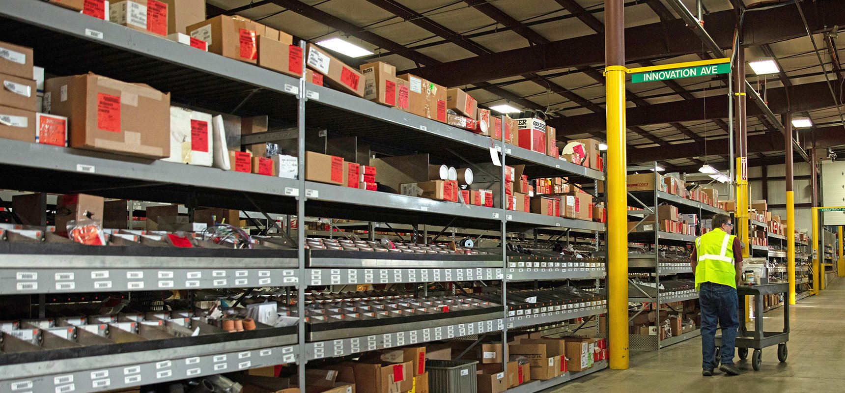 Pierce product support supply warehouse with shelves stocked full of boxes, showing an employee pushing a cart of parts down an aisle. 
