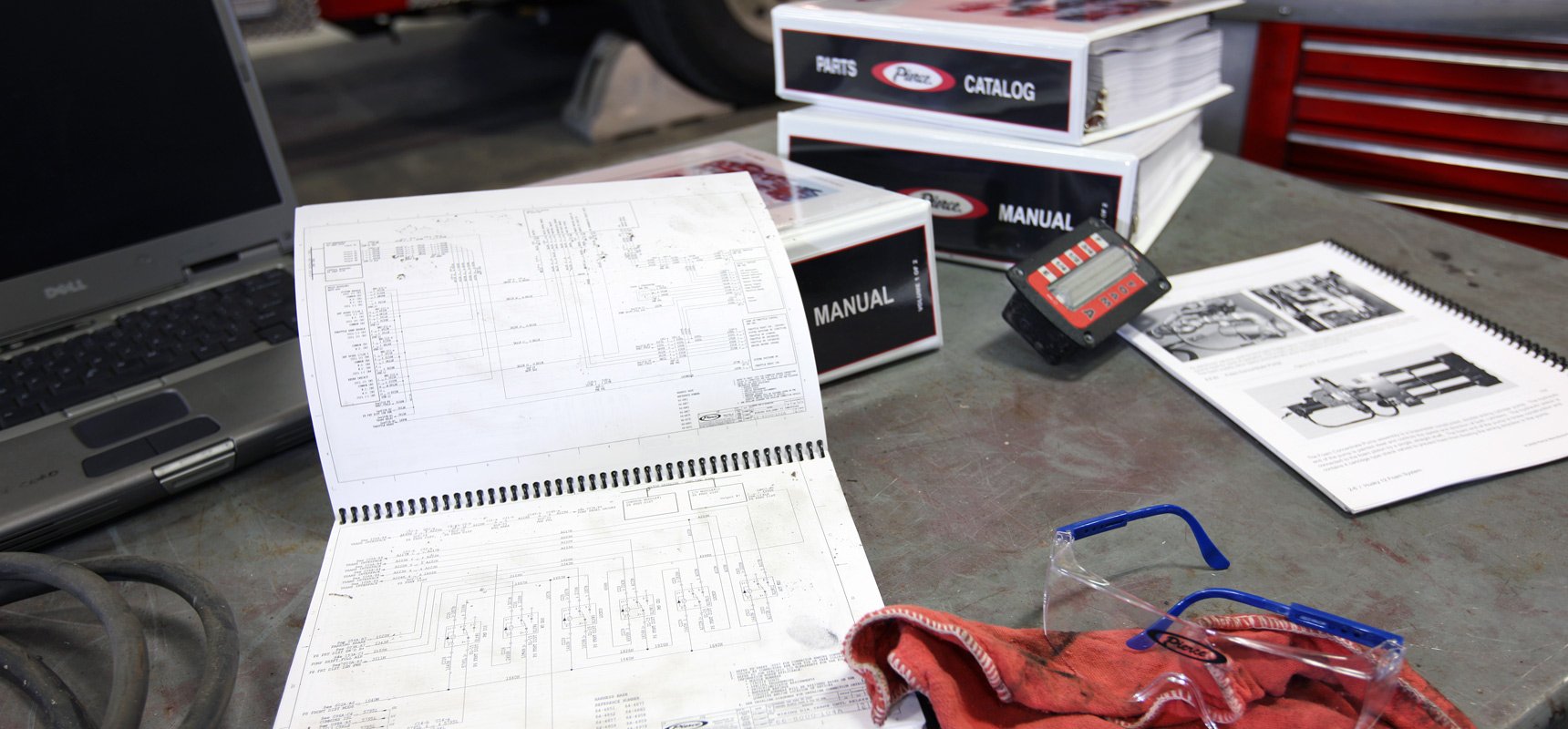 A workbench desk in a Pierce shop floor with a laptop, safety glasses and workbook scattered on top.