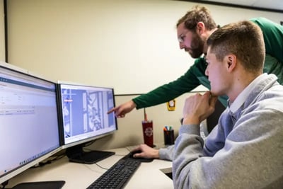 Two engineers review a 3D CAD design of a fire truck on a dual monitor computer setup.