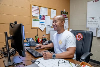 A firefighter sits at a desk reviewing a fire truck purchase order on a computer monitor.