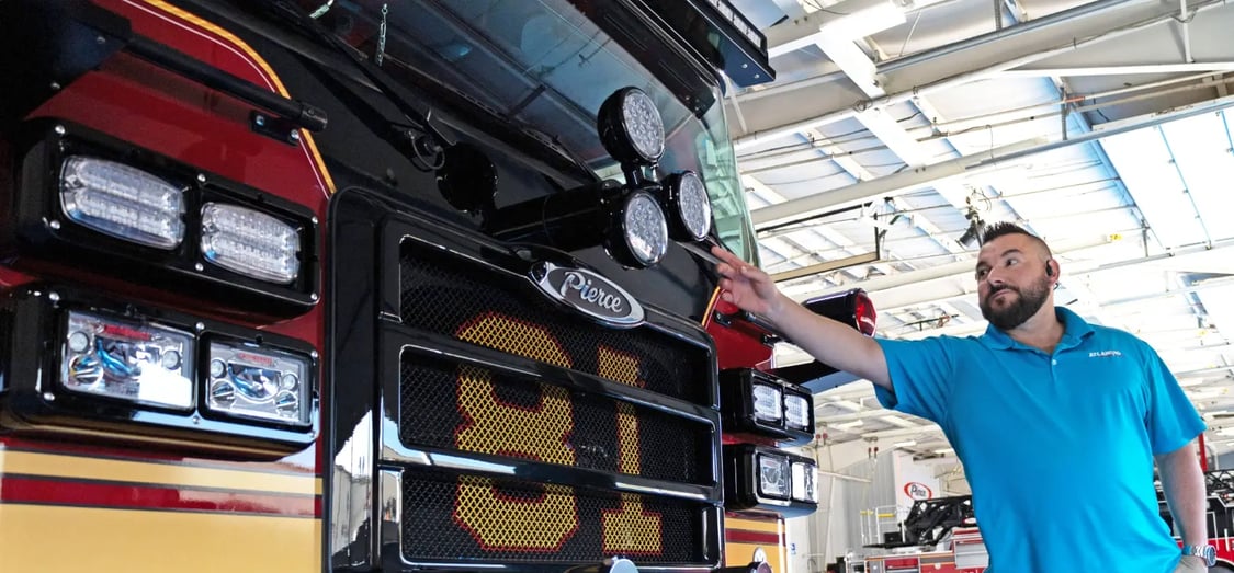 A man in a blue shirt points to the front headlight assembly of a Pierce fire truck during an inspection.