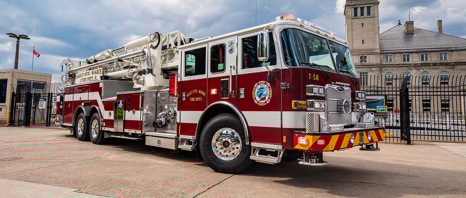 A red and white aerial fire apparatus is parked on an angle facing forward with secured facility in the background.