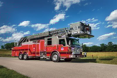 A red and grey aerial fire truck with an elevating platform is driving down a country road with yellow fields, green trees and blue sky in the background.