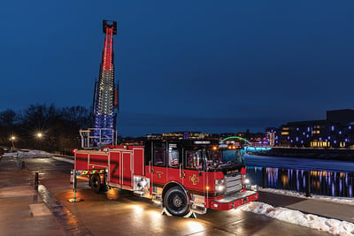 A red and black aerial fire truck is pictured at night next to a river with scene lighting illuminated and the ladder extended showing blue, white and red lighting.