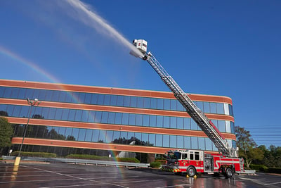 A red and white pumper fire apparatus with an aerial device is pictured with the aerial device deployed shooting water in front of a large glass and brick building.