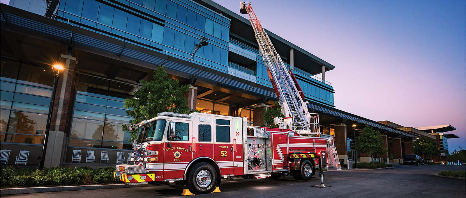 A red and white pumper fire apparatus with an aerial device is pictured with the aerial device deployed shooting water in front of a large glass and brick building.