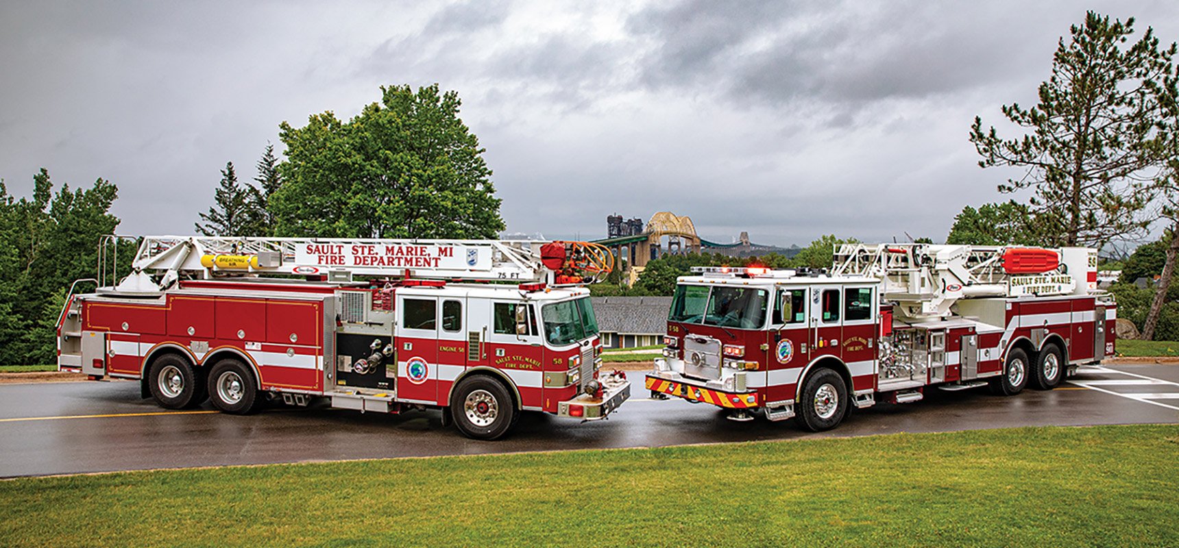 Two red and white aerial fire trucks are parked facing each other with grass in the foreground and a large bridge in the background. 