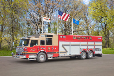 A rescue fire truck is parked on asphalt in front of three flags and an autumn tree colored background. 
