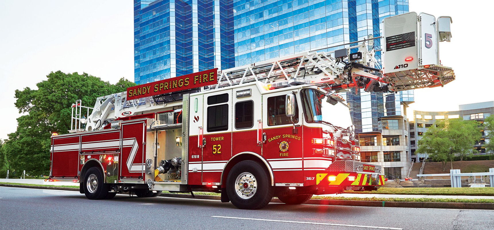 A red and white Pierce quint fire truck is driving on an asphalt road past a mirrored city building.