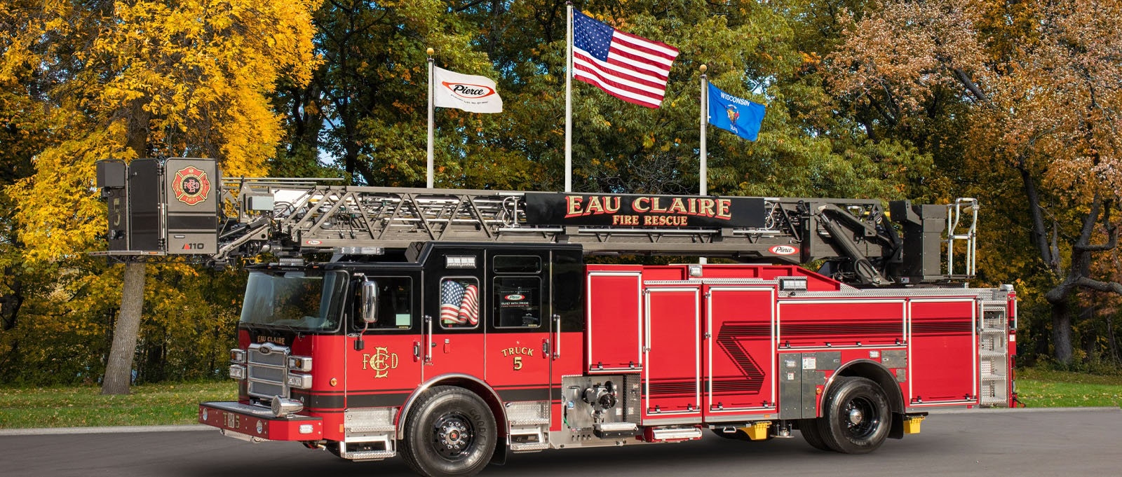 A red and black quint fire truck from Eau Claire Fire Rescue is parked in a lot in front of three flags and autumn-colored trees at Pierce Manufacturing.