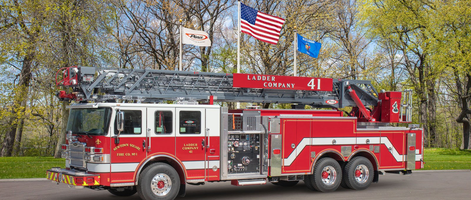 The Newtown Square Fire Company No.1 aerial truck is parked at Pierce Manufacturing in front of three flags and several trees with a blue sky background. 
