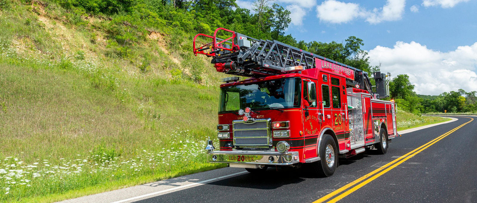 A red quint fire truck with a black stripe drives on an asphalt road next to a green hillside with a blue sky and white clouds in the background. 